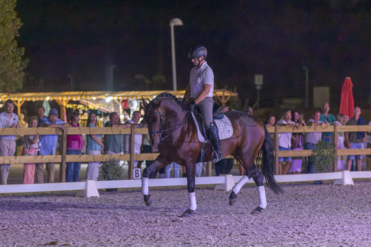 A dark brown horse with white leg wraps is being ridden by a man in equestrian attire inside an outdoor night arena, with spectators visible behind a wooden fence in the background.