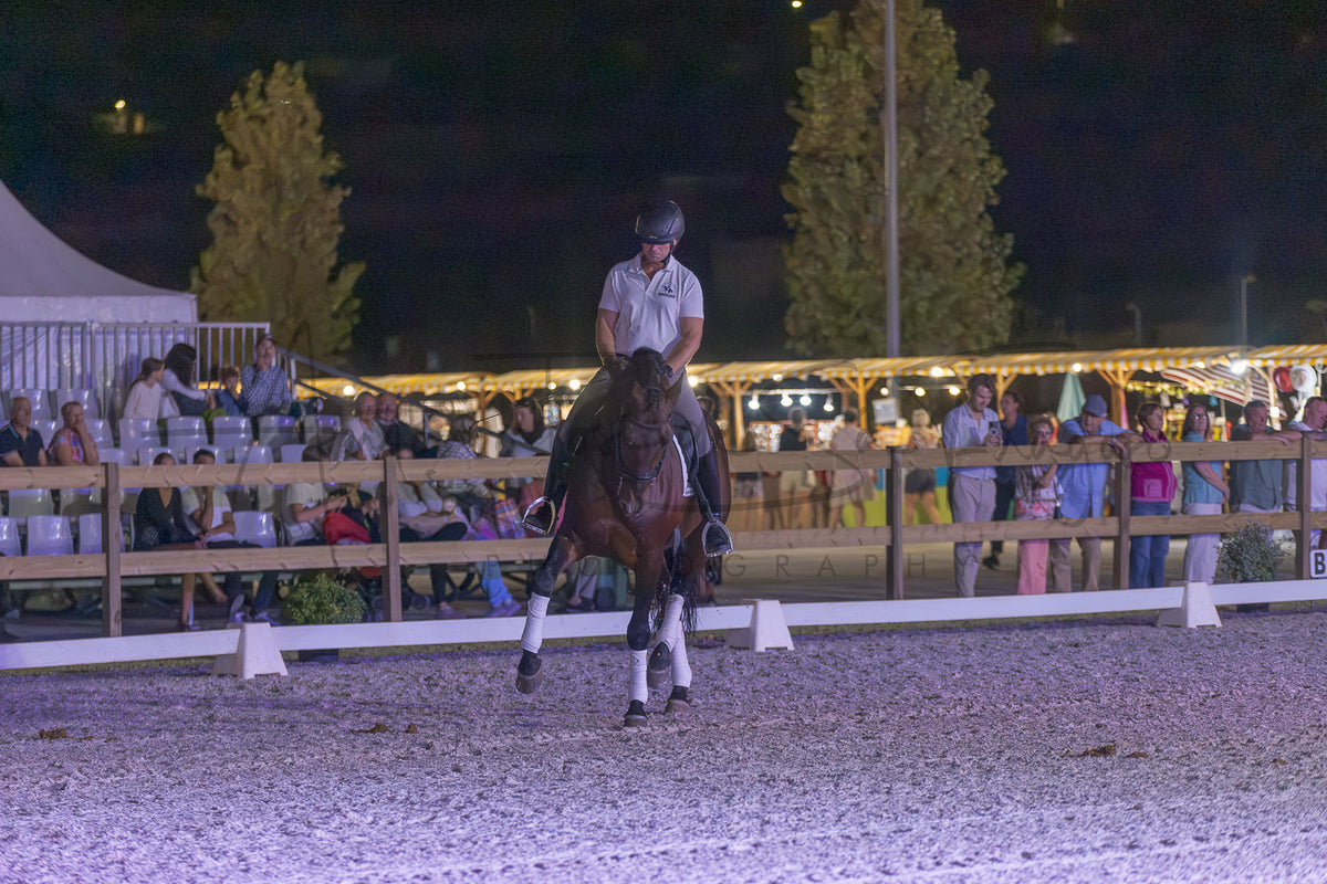 A person riding a dark horse in an outdoor equestrian arena at night, with spectators and tents visible in the background behind a fence.
