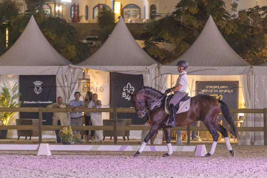 A dark brown horse with white leg wraps is being ridden by a person in formal equestrian attire within an outdoor arena at night. Behind the horse are white event tents with banners, and a few spectators are visible near the fencing.