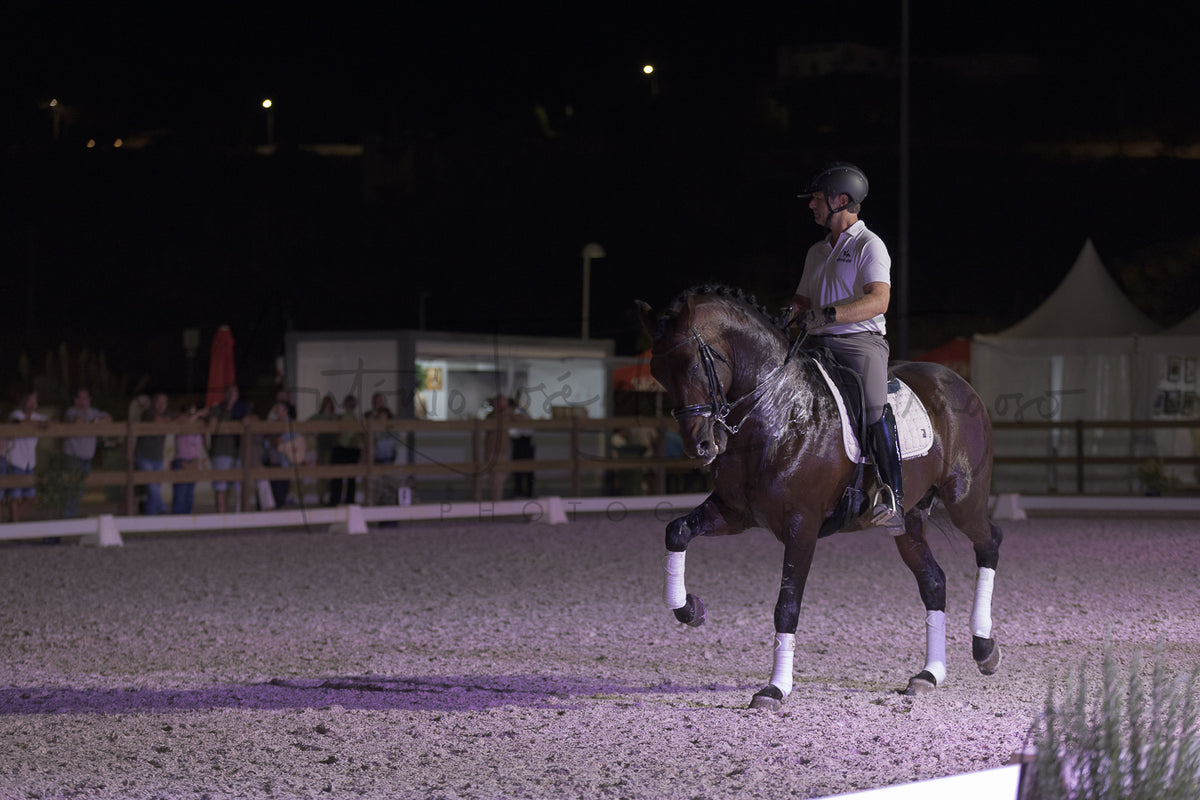 A dark brown horse with white leg wraps is being ridden by a person in equestrian attire inside an outdoor riding arena at night under artificial lighting.