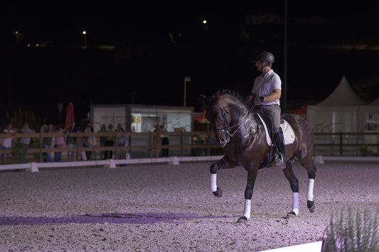 A dark brown horse with white leg wraps is being ridden by a person in equestrian attire inside an outdoor riding arena at night under artificial lighting.
