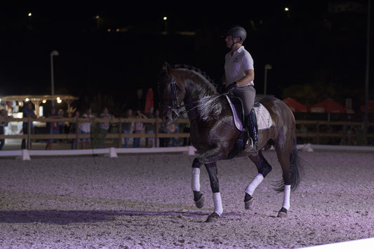 A dark horse with white leg wraps is being ridden by a person in equestrian attire inside an outdoor riding arena at night.