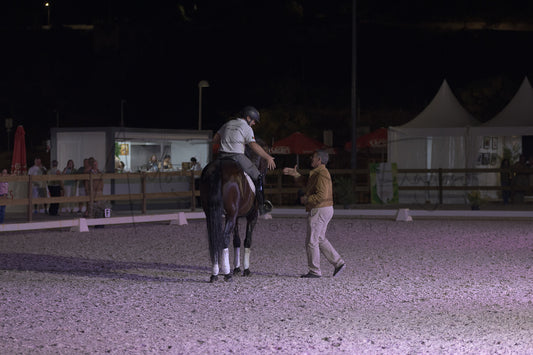 A person in equestrian attire rides a dark horse while another person walks beside them in an outdoor night event arena with tents and spectators visible in the background.
