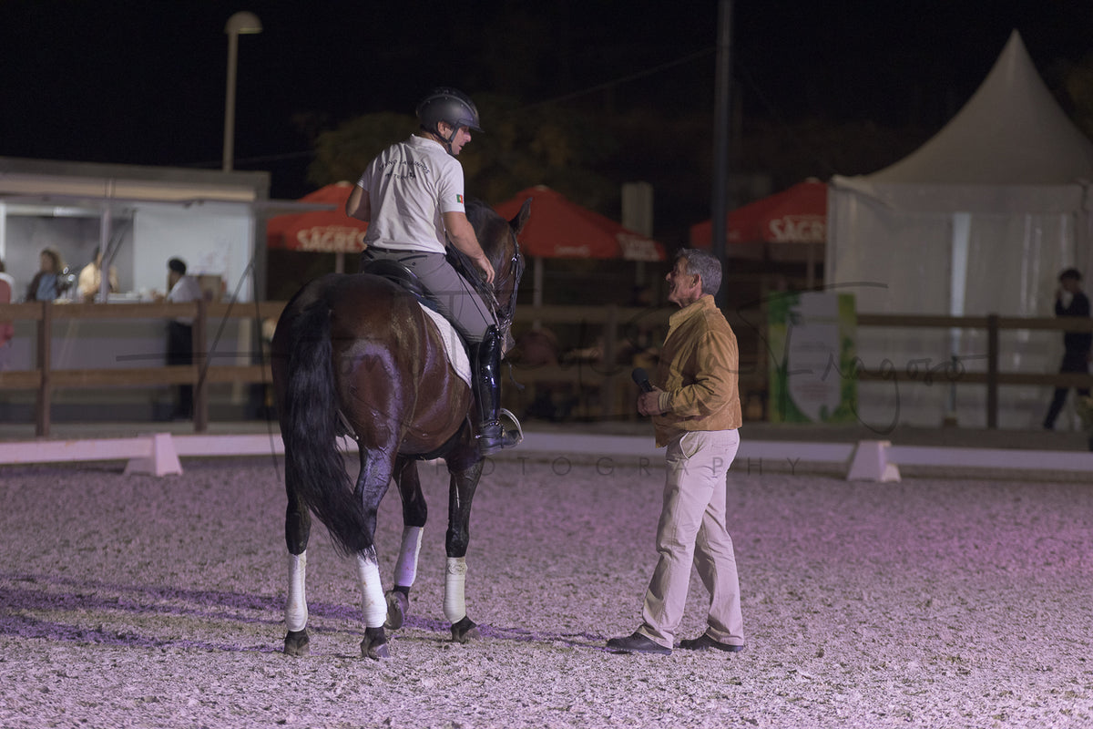 A person in equestrian attire sits on a dark brown horse inside an outdoor riding arena at night, while another man stands nearby. Tents, fencing, and event lighting are visible in the background.