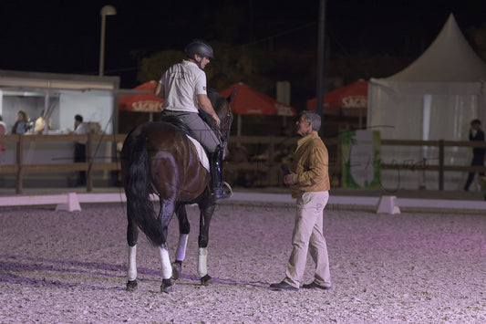 A person in equestrian attire sits on a dark brown horse inside an outdoor riding arena at night, while another man stands nearby. Tents, fencing, and event lighting are visible in the background.