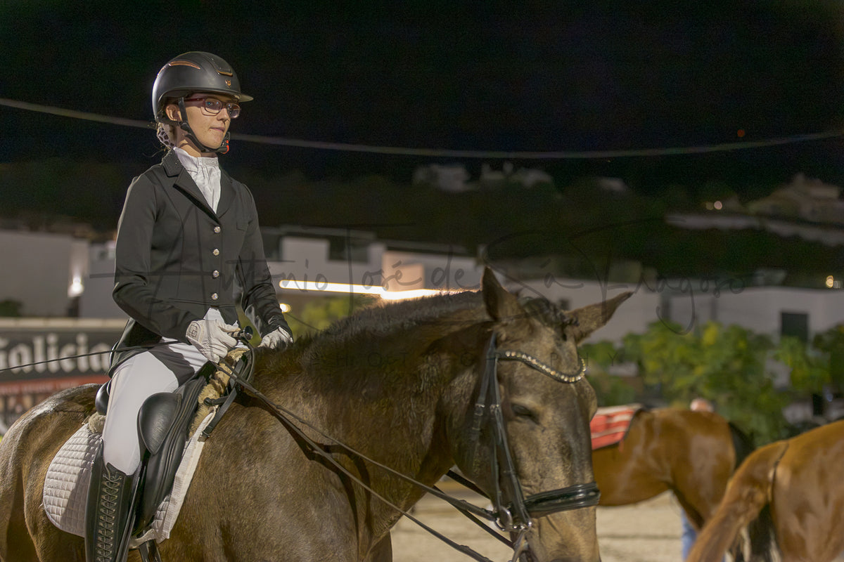 A woman in equestrian attire, including a black helmet, black jacket, white breeches, and gloves, riding a dark brown horse at night in an arena. The photo has a warm, low-light ambiance with a watermark across the center.