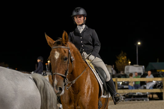 A rider in formal equestrian attire sits on a brown horse in a night-time outdoor arena, with a dark sky and distant lights in the background.