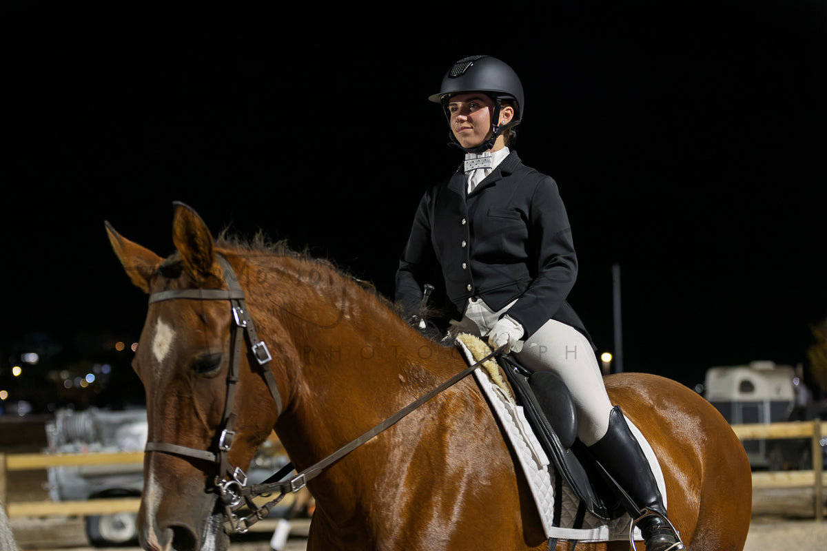 A rider in traditional equestrian attire sits on a brown horse at night in an arena. The rider wears a black helmet, black jacket, white breeches, and black boots, while the horse has a glossy brown coat and a white patch on its forehead.
