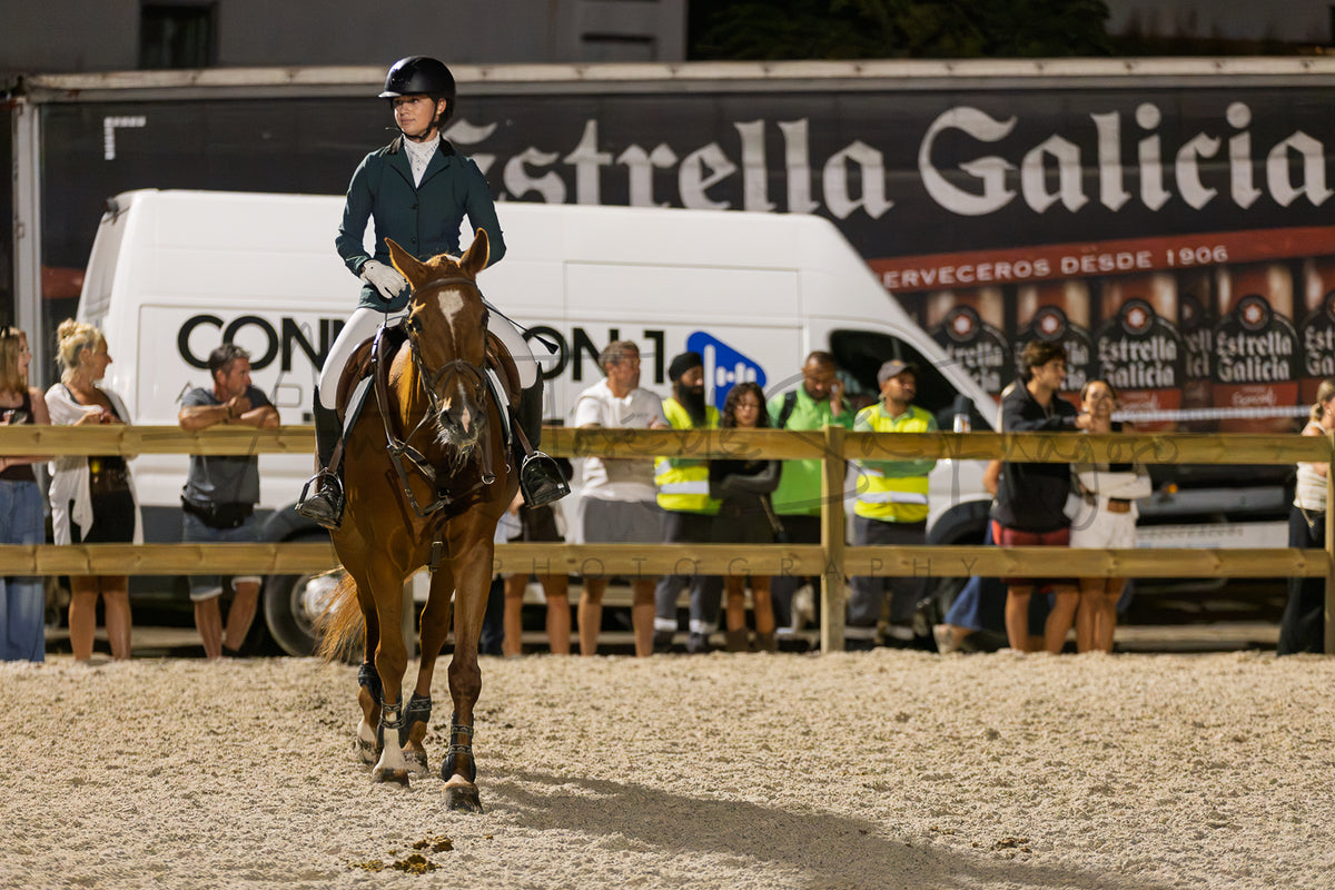 A horse rider in formal equestrian attire rides a brown horse in a sandy arena, with spectators and a white van behind a wooden fence. A large Estrella Galicia banner is visible in the background.