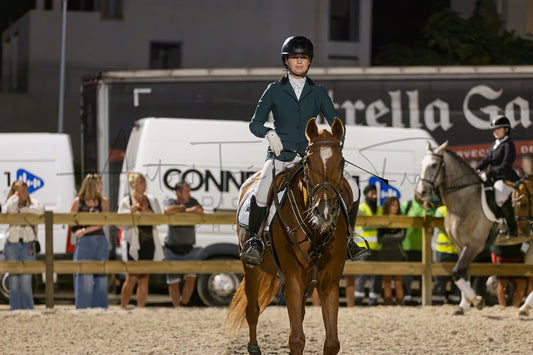 A rider in formal equestrian attire sits on a brown horse in an indoor arena, with spectators and other horses visible in the background at night.