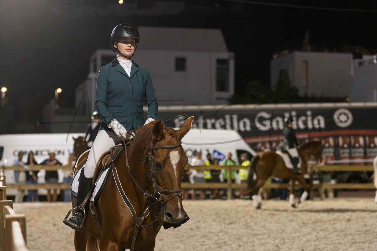 A person in equestrian attire riding a brown horse at night in an outdoor arena, with other horses and event structures blurred in the background.