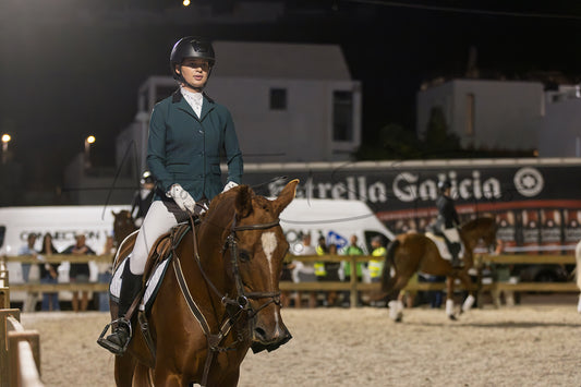 A person in equestrian attire riding a brown horse at night in an outdoor arena, with other horses and event structures blurred in the background.