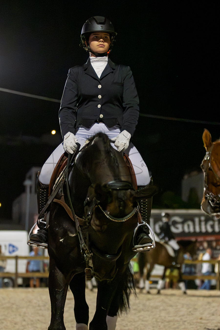 A person in formal equestrian attire sits on a dark horse in an indoor riding arena at night, wearing a black helmet, dark jacket, white breeches, gloves, and tall boots. Another horse is partially visible at the right edge, and arena lighting illuminates the scene.
