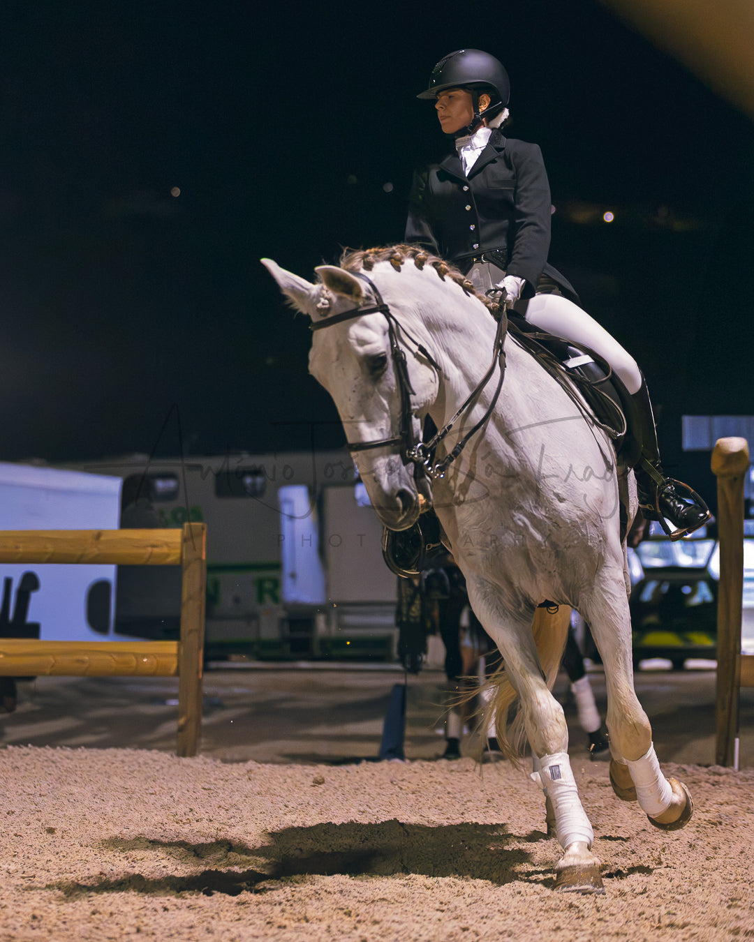 A rider in a black equestrian jacket, white breeches, and a black helmet rides a white horse during a night competition in an indoor arena.