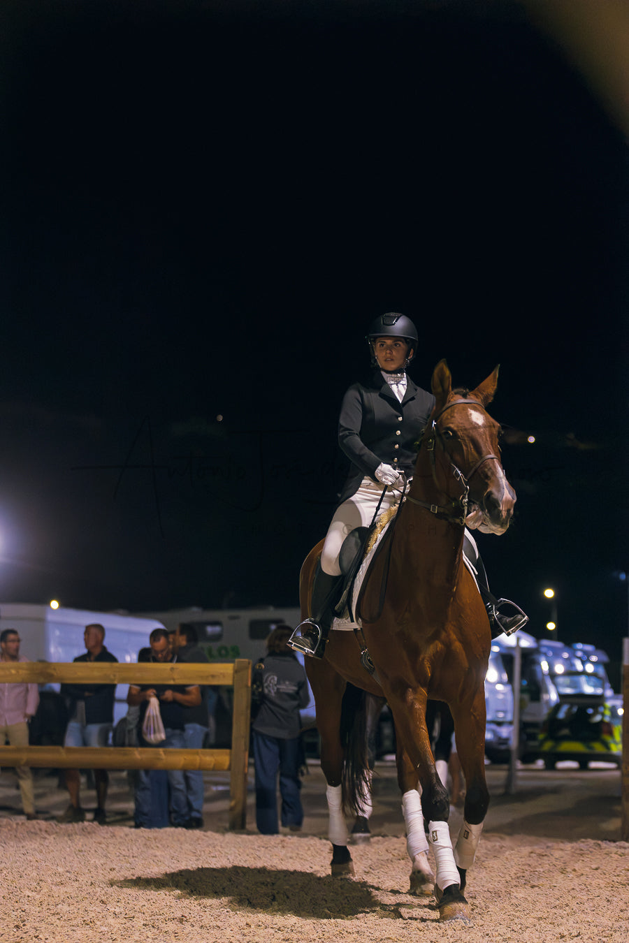 A rider in formal equestrian attire sits on a brown horse in an outdoor arena at night, with spectators and vehicles visible in the background.