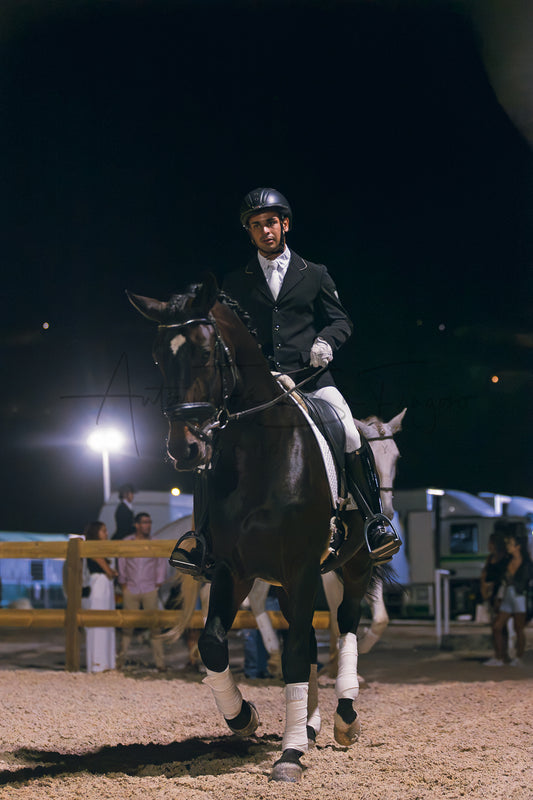 A man in formal equestrian attire rides a dark brown horse in an indoor arena at night, with bright arena lights and spectators visible in the background.