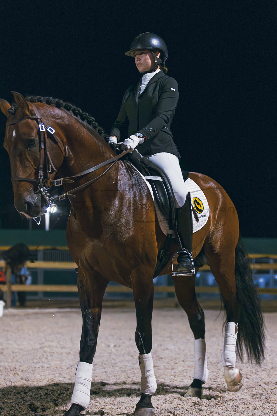 A woman in equestrian riding attire sits on a brown horse inside an indoor riding arena at night. The rider wears a black helmet, dark jacket, white breeches, and gloves, while the horse has white leg wraps and a black mane.