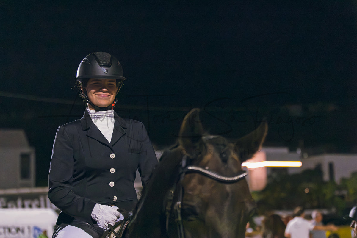 A woman in equestrian attire, including a black helmet, dark jacket, white shirt, and gloves, riding a dark horse at night in an outdoor setting.