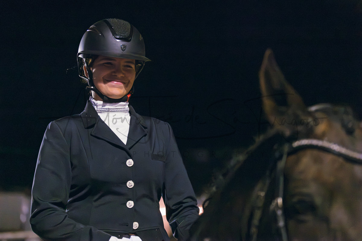 A woman in equestrian attire, wearing a black riding helmet and a black riding jacket over a white shirt, seated on a horse against a dark background.
