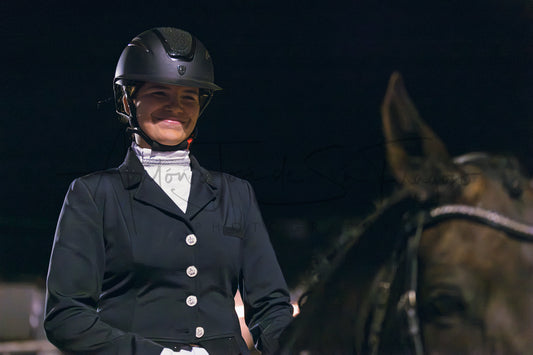 A woman in equestrian attire, wearing a black riding helmet and a black riding jacket over a white shirt, seated on a horse against a dark background.