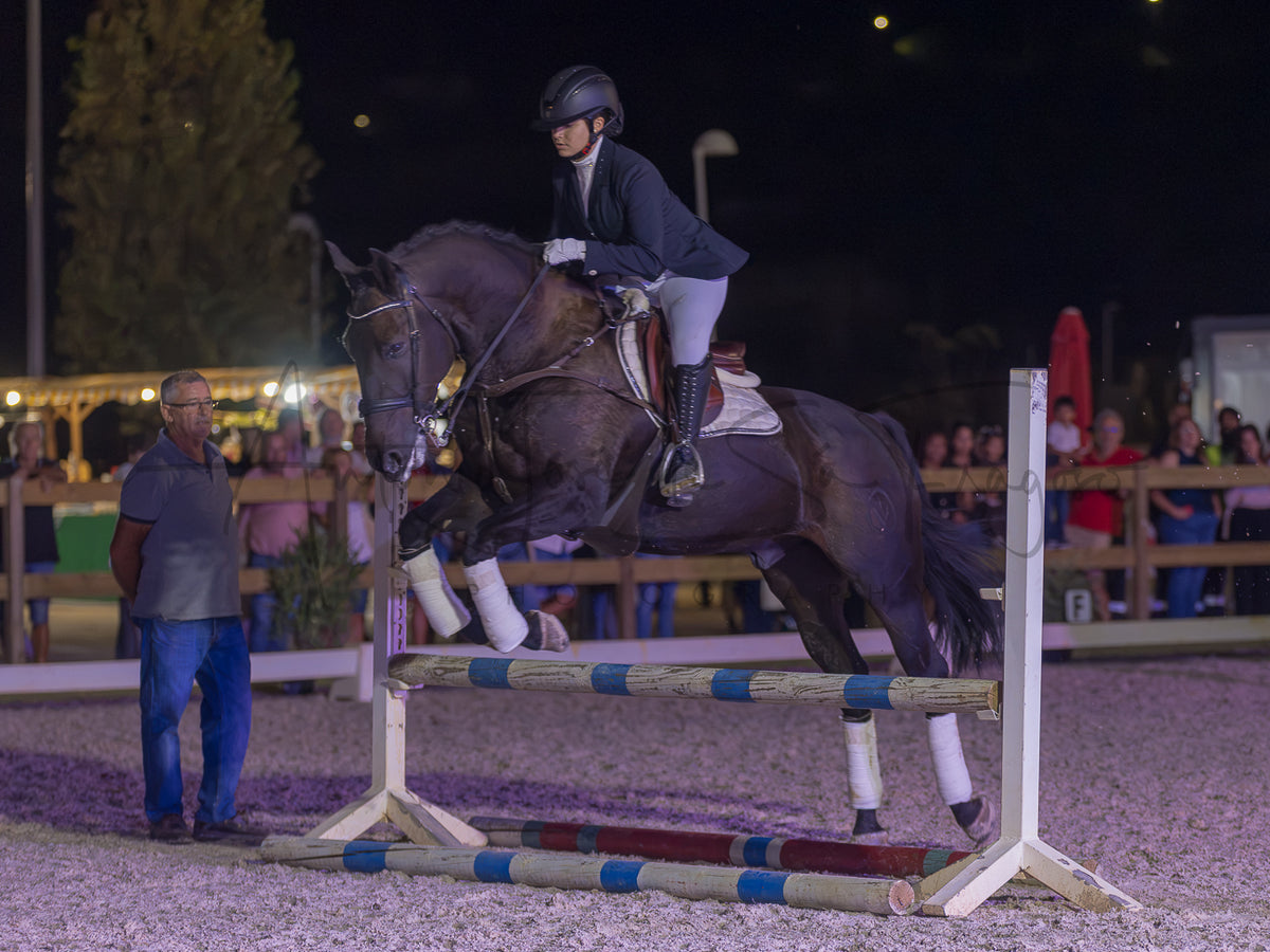 A horse rider on a dark horse jumps an equestrian obstacle in an outdoor night arena, with spectators and a man standing nearby.