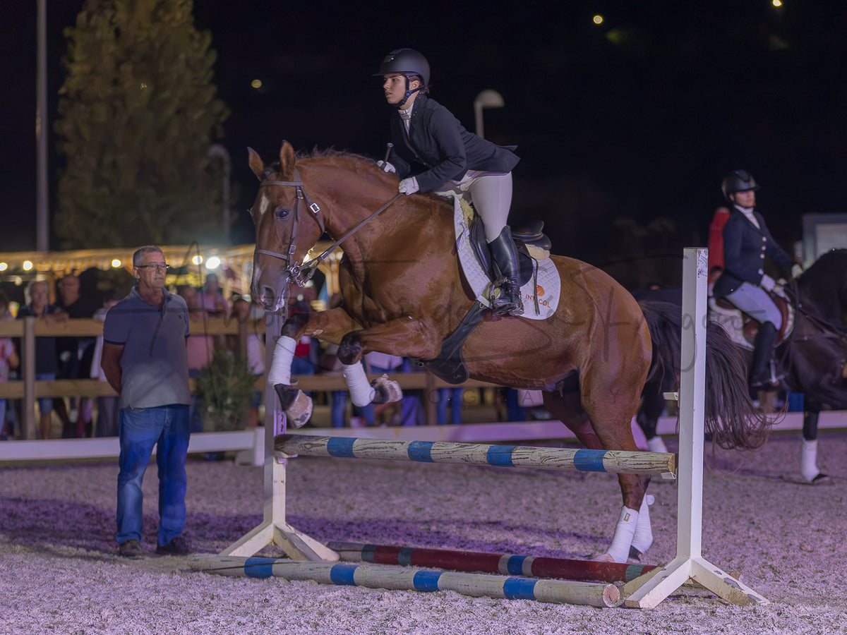 A brown horse with white leg wraps is being ridden by a person in equestrian attire while jumping a striped obstacle in an outdoor night equestrian arena. Another rider is visible in the background.