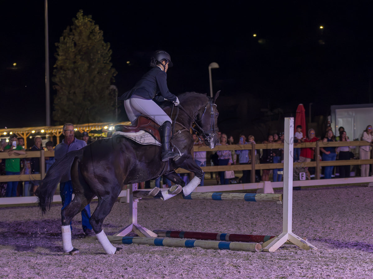 A dark horse with white leg wraps is being ridden by a person in equestrian attire while jumping an obstacle in an outdoor night arena with spectators in the background.