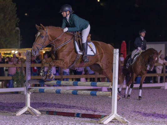 A horse rider in a green jacket and white breeches jumps a horse over a striped obstacle in an equestrian arena at night, with another rider and spectators blurred in the background.