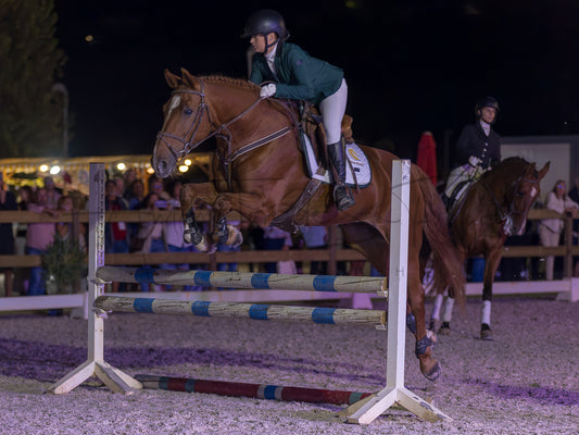 A horse show jumping competition at night in an arena, with a rider on a brown horse mid-jump over a striped obstacle, and another rider and horse in the background.
