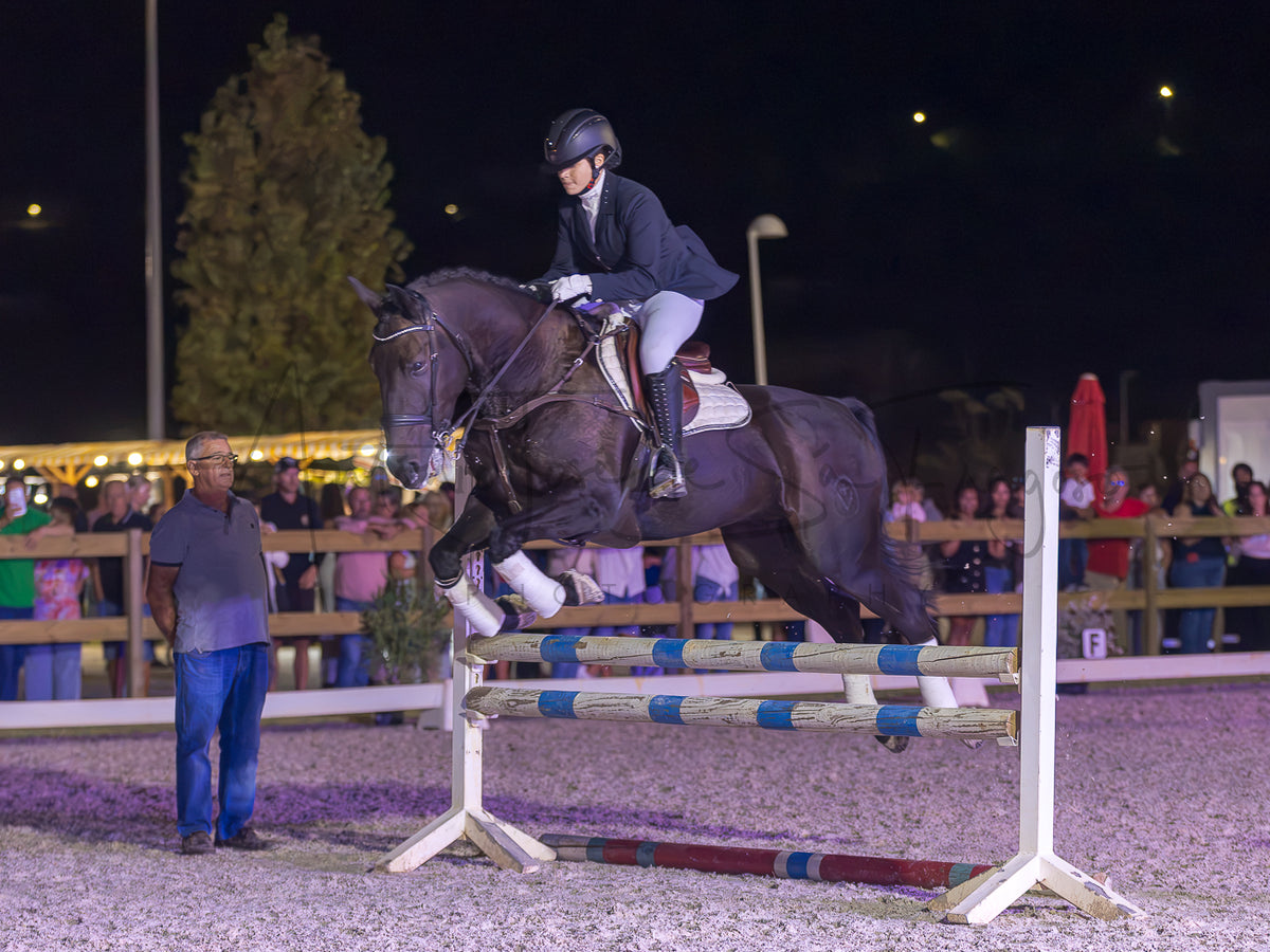 A dark brown horse with white socks is being ridden by a person in equestrian attire as it jumps over a striped obstacle in an outdoor night equestrian arena, with spectators and a judge visible in the background.