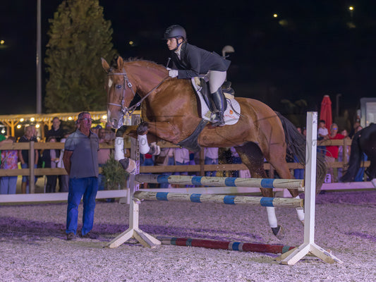A horse rider on a brown horse jumps an obstacle in an outdoor night equestrian arena, with spectators and lighting visible in the background.