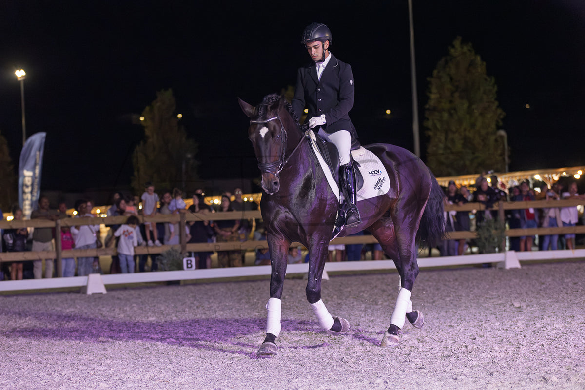 A dark horse with white leg wraps is being ridden by a person in formal equestrian attire inside an outdoor arena at night, with spectators visible in the background.