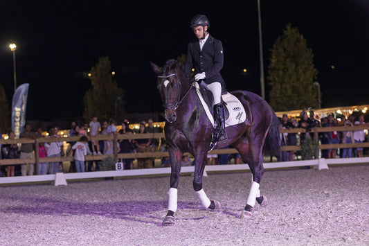 A dark horse with white leg wraps is being ridden by a person in formal equestrian attire inside an outdoor arena at night, with spectators visible in the background.