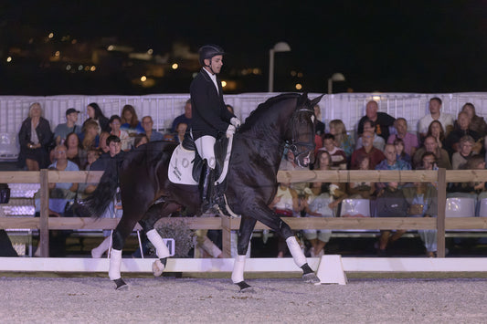 A rider in a black equestrian jacket and white breeches rides a dark horse with white leg wraps around an outdoor arena at night, with spectators seated in the background.