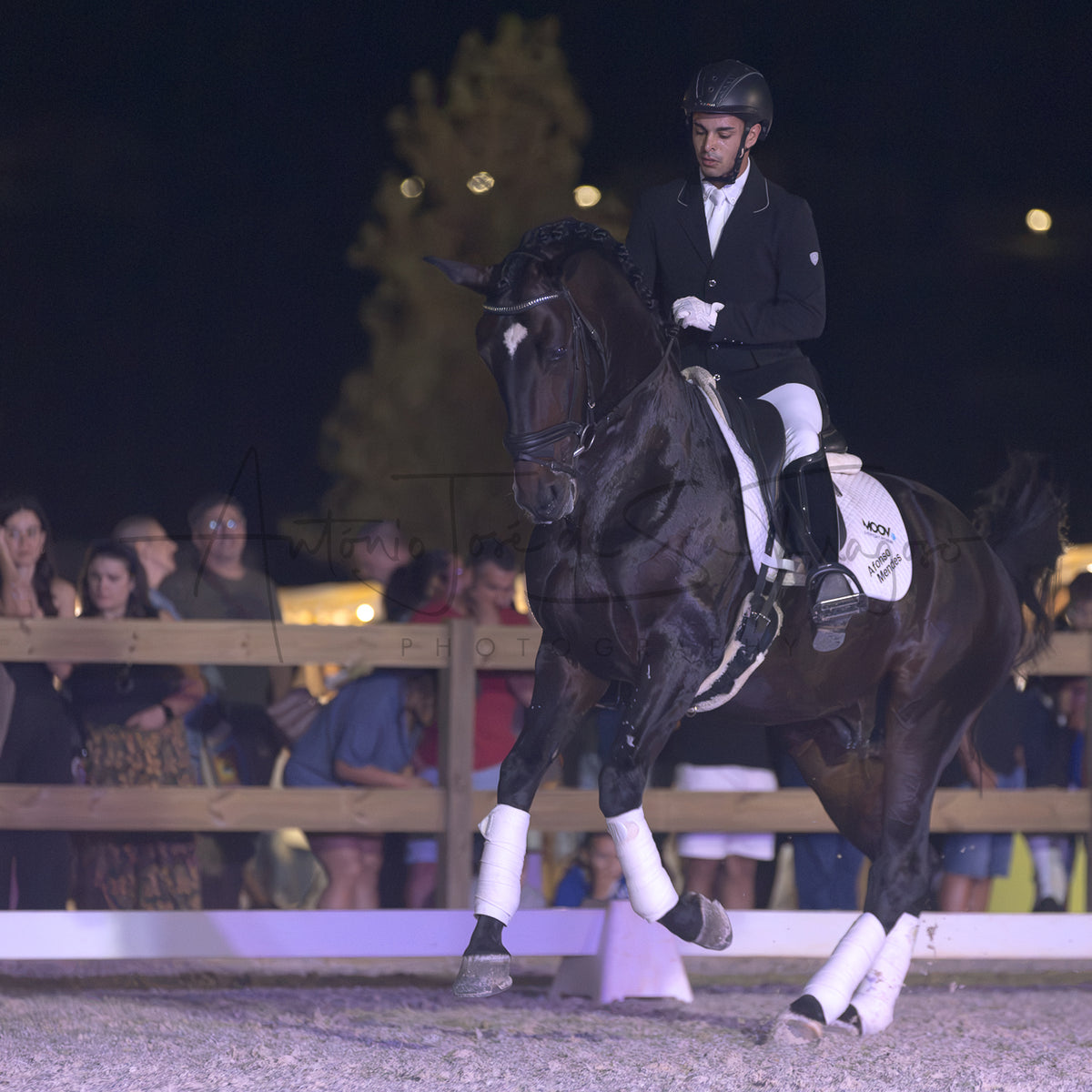 A dark brown horse being ridden by a male equestrian in formal riding attire, photographed at night in an arena with spectators visible behind a fence.