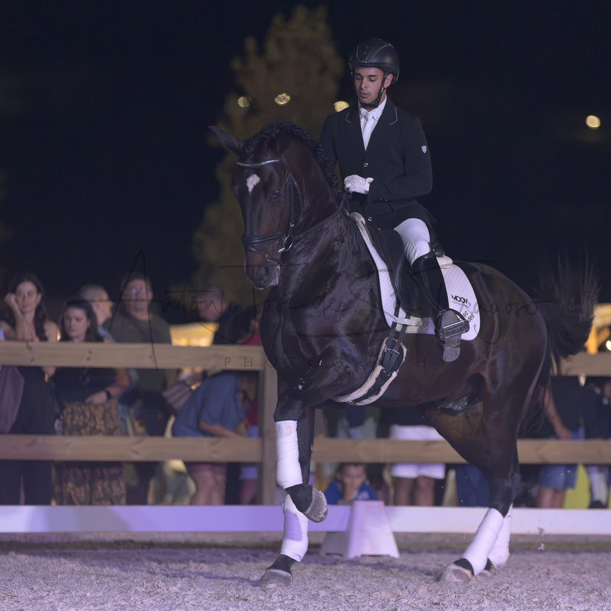 A dark horse with white leg wraps is being ridden by a person in formal equestrian attire inside an outdoor arena at night, with spectators visible behind a fence in the background.
