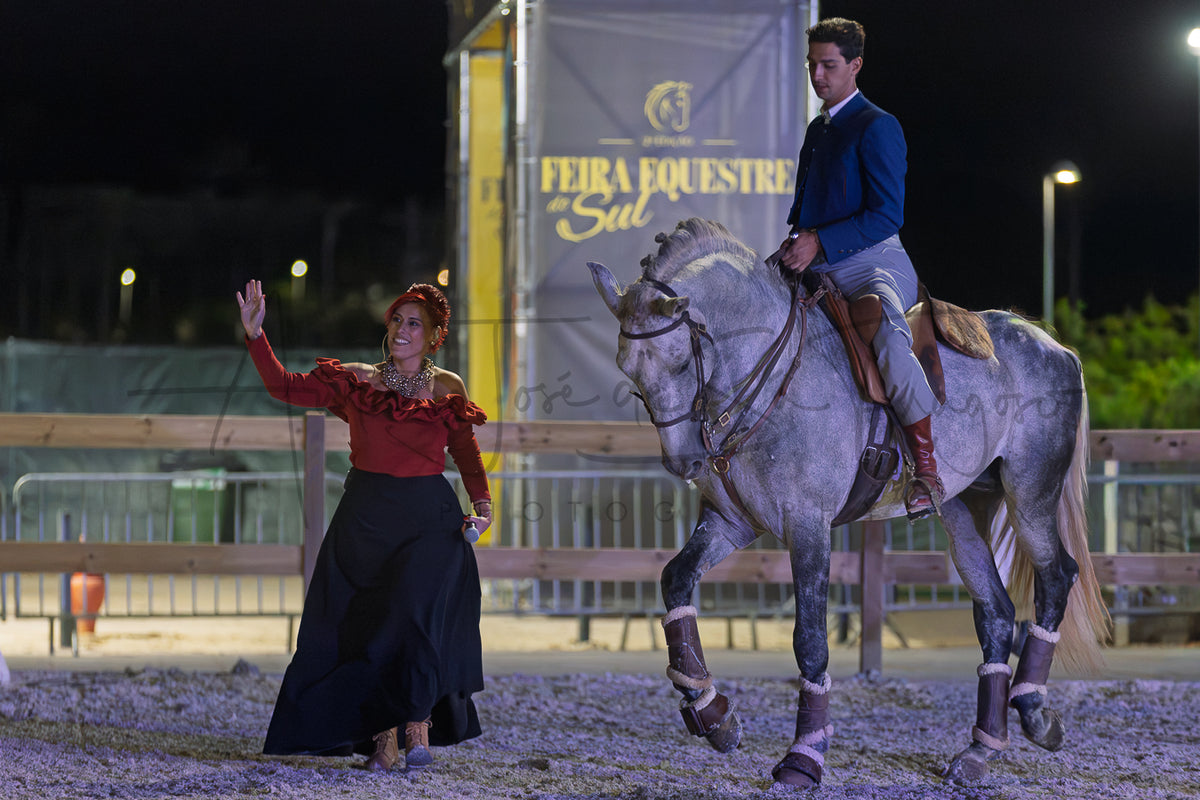 A man in formal equestrian Portuguese Traditional attire rides a dappled gray horse at night in an arena, while a woman in a red ruffled dress waves beside him. A large banner reading 'FEIRA EQUESTRE Sul' is visible in the background.