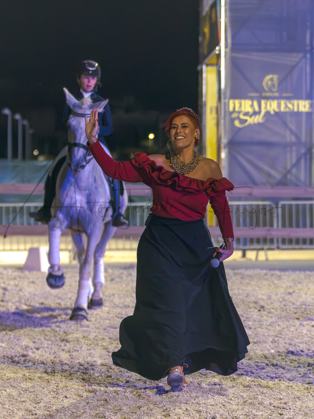 A woman in a red off-the-shoulder ruffled top and a long black skirt waves to the camera while walking in an equestrian arena at night. A rider on a horse is visible in the background, along with a banner reading 'FEIRA EQUESTRE Sul'.