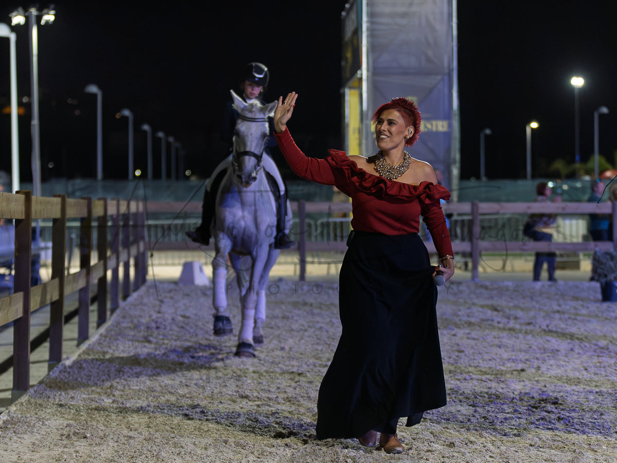 A woman in a red ruffled off-the-shoulder top and a long black skirt waves to a rider on a white horse inside a lit equestrian arena at night.