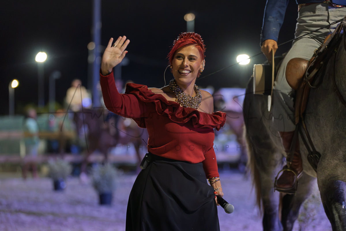 A woman in a red ruffled off-the-shoulder top and black skirt waves to a crowd at night, standing in an outdoor arena next to a horse.