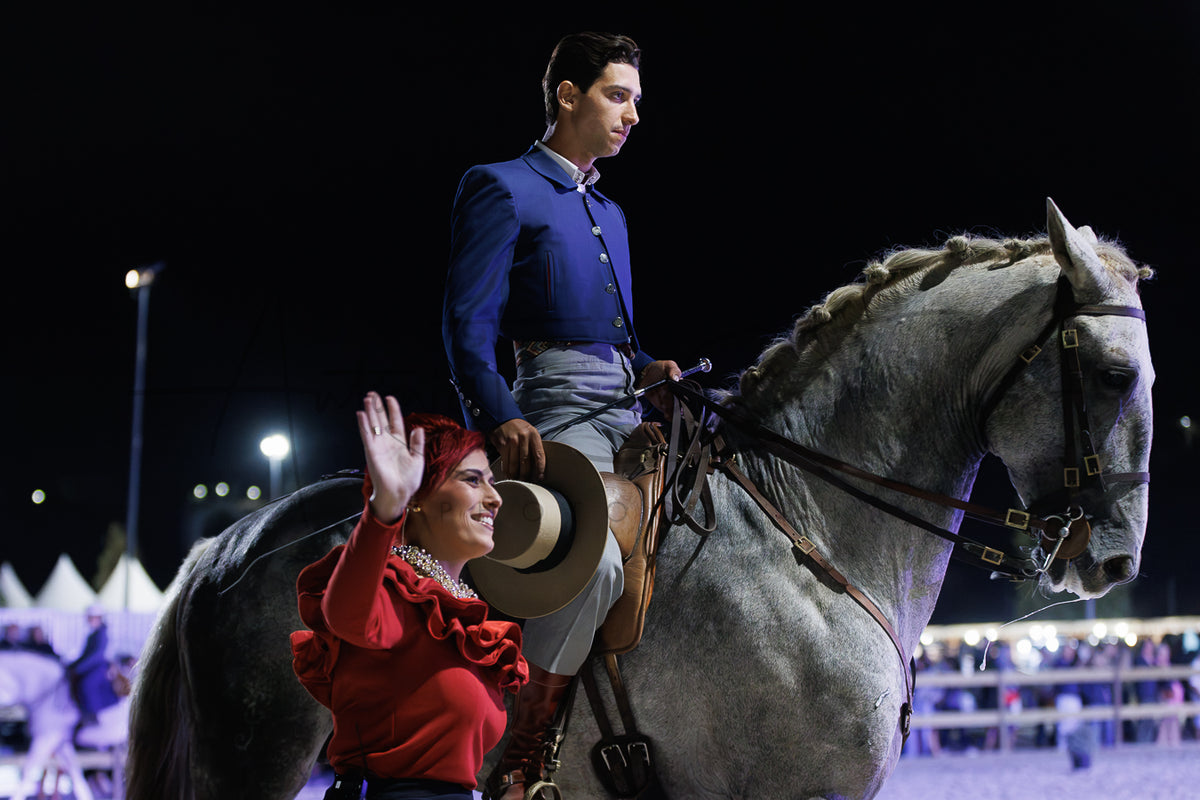 A man in a blue mariachi jacket and gray pants rides a large gray horse at night while a woman in a red top and brown hat waves to the crowd. The scene is photographed in an outdoor arena with stadium lights and tents visible in the background.