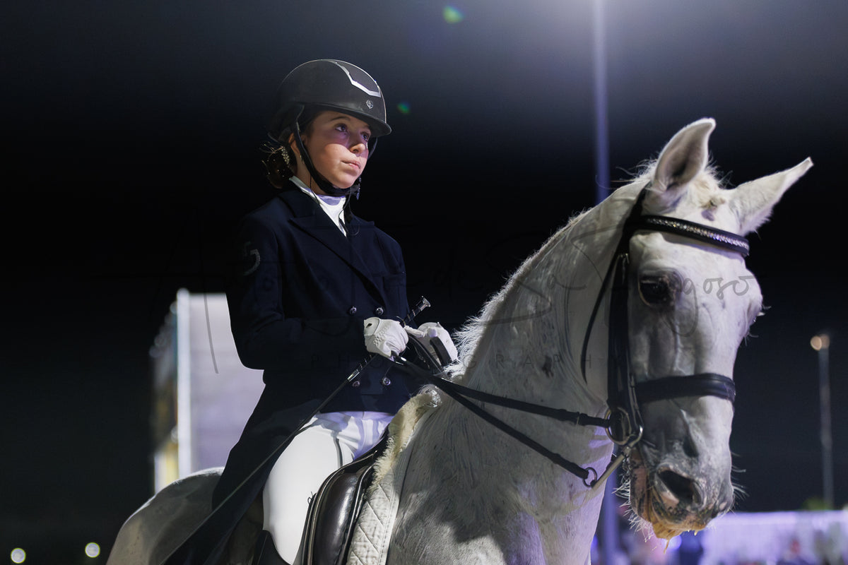 A woman in equestrian attire, wearing a black helmet, navy jacket, white breeches, and gloves, riding a white horse at night under bright stadium lights.