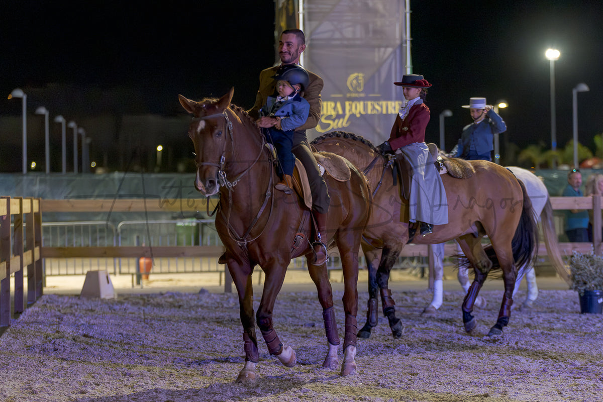 A man rides a brown horse in an equestrian arena at night while holding a child, with other riders and horses visible behind him. A large banner in the background reads 'Feira Equestre do Sul'.