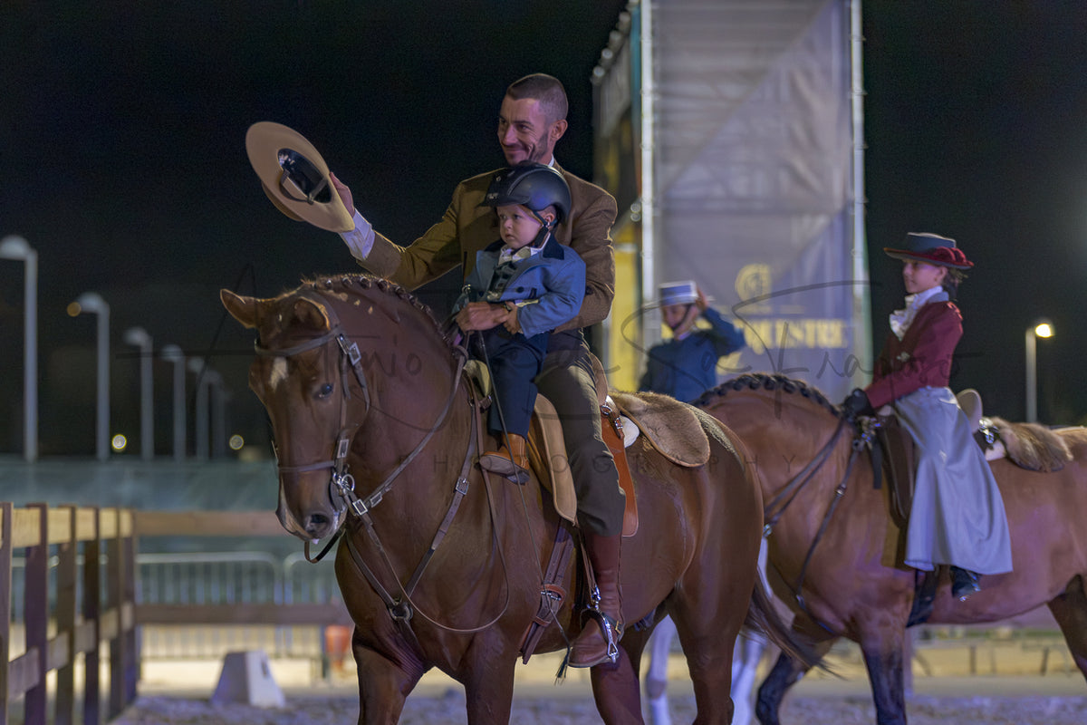A man in historical Portuguese Traditional attire rides a brown horse at night while holding a child, with two young girls in period costumes riding horses beside them on a track.