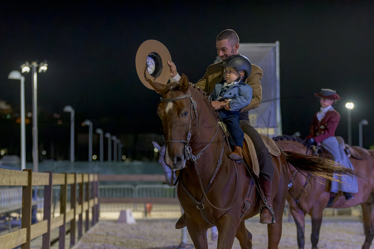 A man rides a brown horse at night in an equestrian arena, wearing a cowboy hat and holding a child. Another rider is visible in the background under stadium lights.