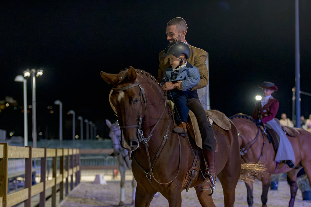 A man rides a brown horse at night while holding a young child on his lap. Another rider is visible in the background on a horse in an outdoor arena with stadium lights on.