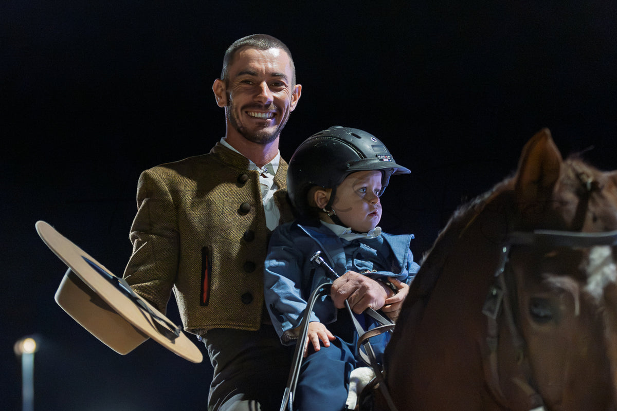 A man in a brown traditional jacket smiles while riding a horse with a young child in a blue equestrian suit and black helmet. The man holds a beige Portuguese Traditional hat in one hand against a dark background.