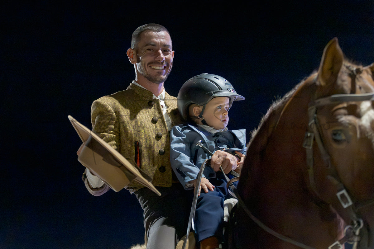 A man in a gold equestrian jacket smiles while holding a Portuguese Traditional hat, standing beside a child in riding attire and a helmet on a brown horse against a dark background.