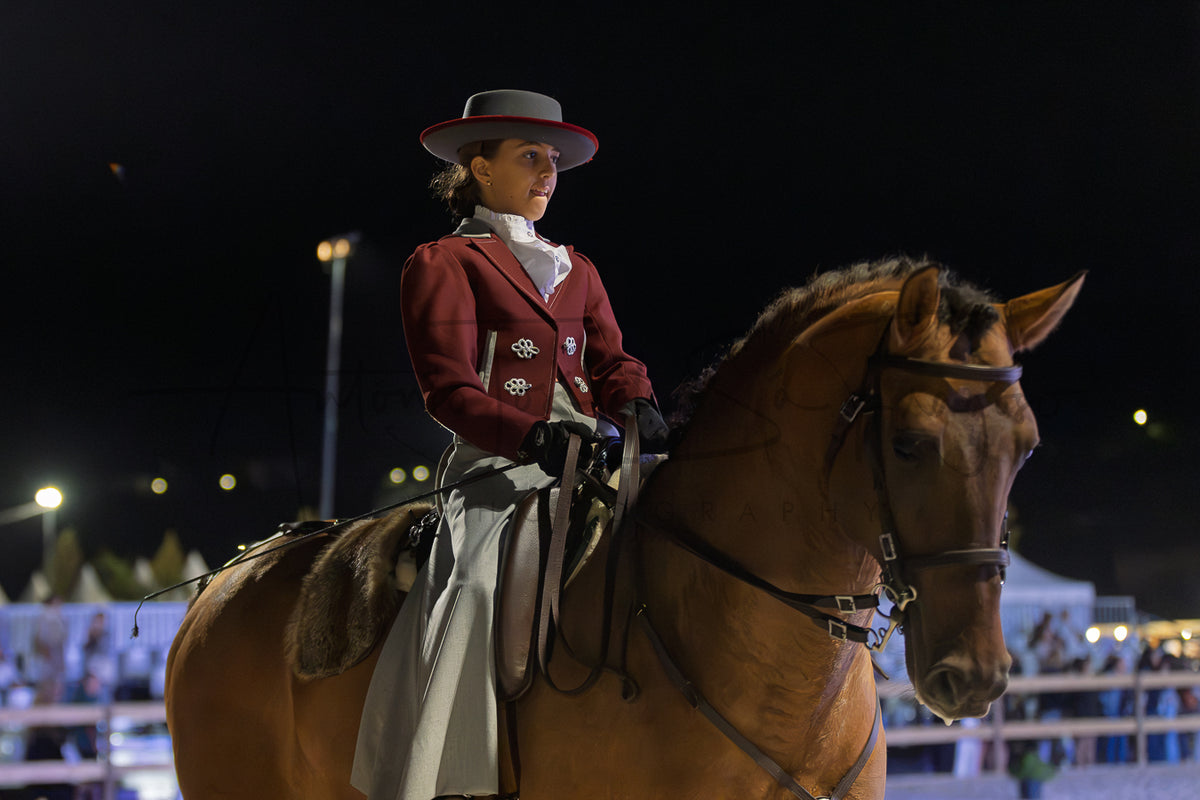 A woman in formal equestrian Portuguese Traditional attire, including a dark red jacket, white skirt, and gray hat, riding a brown horse at night in an arena.
