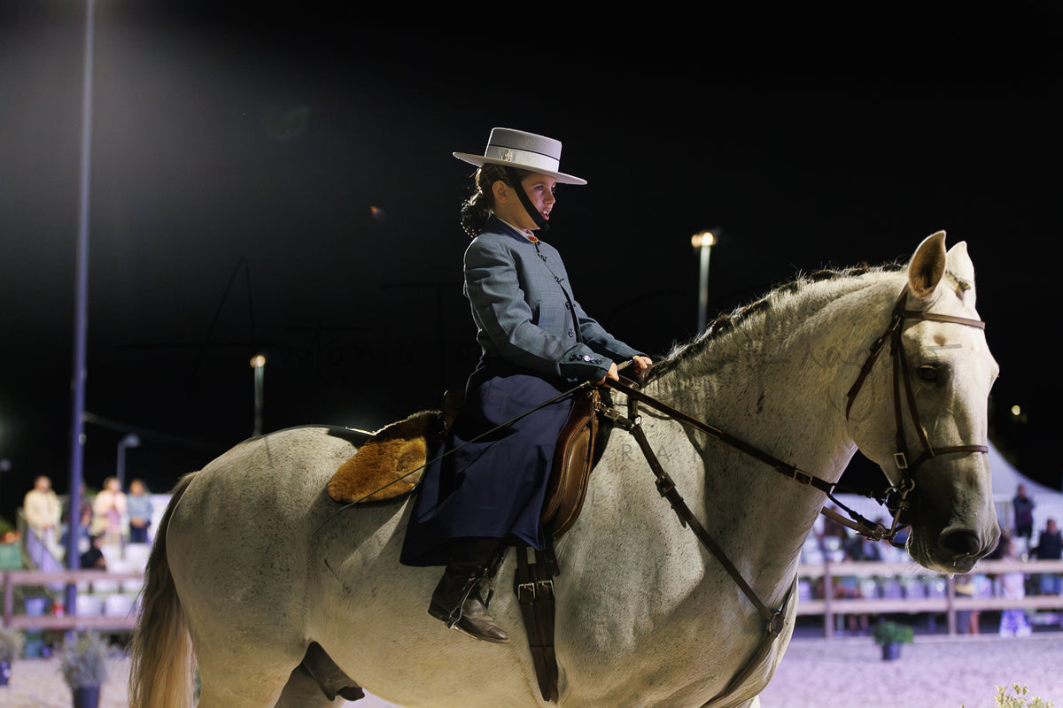 A person in a Portuguese Traditional hat and dark western outfit rides a light-colored horse in a night arena with stadium lights and blurred spectators in the background.
